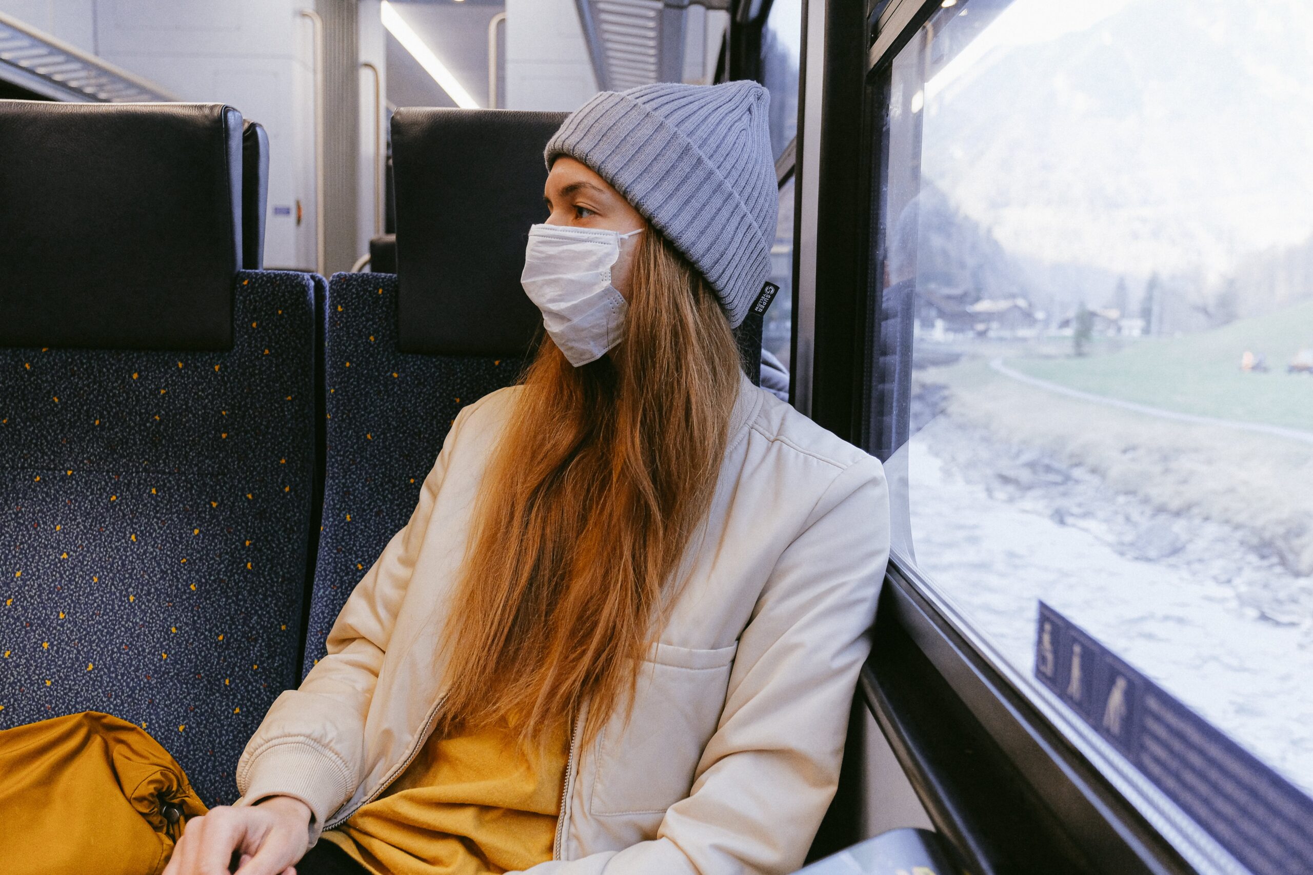 Girl sitting on train with a face mask and woolly hat.